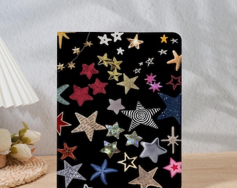 Può includere: Una copertina di taccuino nera adornata da una varietà di stelle in vari colori e trame. Le stelle includono rosso, oro, argento e blu, creando un tema celeste. La copertina è rettangolare e sembra essere realizzata in un materiale resistente.