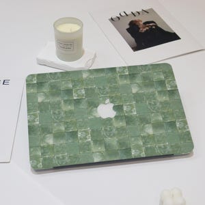 May include: A green laptop case with a square tile pattern and a white Apple logo. A lit candle in a glass jar sits on a white stone, and a magazine is in the background.