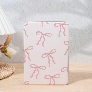 May include: A rectangular, light pink case featuring a repeating pattern of pink bows. The bows are drawn with delicate lines, creating a soft and feminine aesthetic. The case is likely for a phone or tablet.
