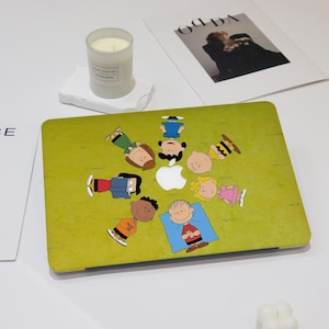 Op de afbeelding: Een groene laptophoes met een cartoonillustratie van personages die op een grasveld liggen rond een wit Apple-logo. De hoes heeft een matte afwerking. Een brandende kaars in een glazen pot staat op een witte standaard.