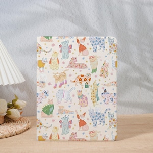 Könnte beinhalten: Eine cremefarbene Tablet-Hülle mit einem skurrilen Muster aus illustrierten Katzen in verschiedenen Posen und Outfits. Die Katzen sind in einer Vielzahl von Farben, darunter Rosa, Blau, Gelb und Grün. Die Hülle ist rechteckig und scheint aus einem strapazierfähigen Material zu sein.