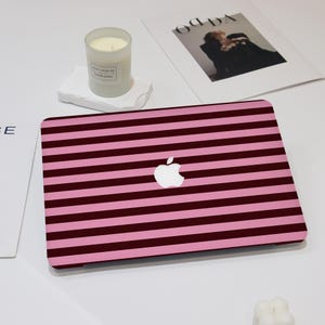 May include: A pink and burgundy striped laptop case with a white Apple logo. A lit candle in a glass jar sits on a white stone, with a magazine in the background.
