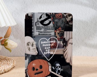 Op de afbeelding: Een rechthoekig decoratief item met Halloween-thema met een collage van griezelige afbeeldingen. Het ontwerp bevat een Ghostbusters-afbeelding, schedels, pompoenen en een spook. De tekst luidt "Surviving purely out of spite."