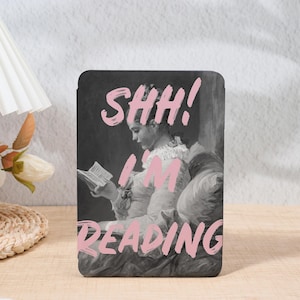 May include: A black and white image of a woman reading a book, with the text "SHH! I'M READING" in pink. The woman is wearing a dress and seated on a cushioned surface. The image is likely a decorative item.