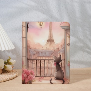 Könnte beinhalten: Notizbuch mit einer Pariser Szene. Eine schwarze Katze sitzt auf einem Balkon mit Blick auf den Eiffelturm. Die Illustration verwendet eine weiche Pastellpalette mit Rosa- und Pfirsichtönen. Ein Strauß rosa Rosen befindet sich im Vordergrund.