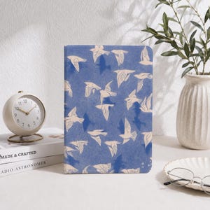 May include: A rectangular notebook with a blue cover featuring a repeating pattern of cream-colored birds in flight. The cover has a textured appearance, and the birds are depicted in various positions, creating a dynamic visual effect across the surface.