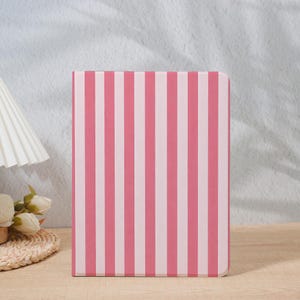 May include: A pink and white striped notebook stands upright on a wooden surface. The notebook's cover features vertical stripes of alternating pink and white. The background includes a white lamp and a decorative floral arrangement.