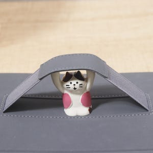 May include: A gray carrying case with a matching handle and a small, white cat figurine with black and pink markings. The cat is holding up the handle of the case.