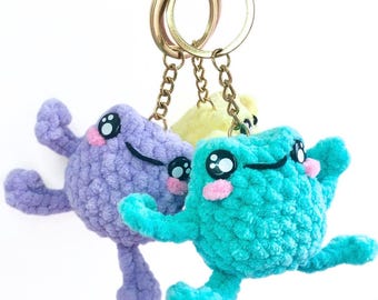 May include: Three handmade crochet frog keychains in purple, yellow, and teal. Each frog has large black eyes, pink cheeks, and is attached to a gold-colored keychain ring. The frogs are in a playful pose.