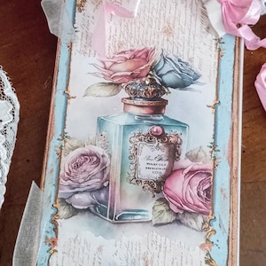 Puede incluir: Una tarjeta decorativa rectangular con una ilustración de estilo vintage de un frasco de perfume rodeado de rosas rosas y azules. El diseño incluye texto manuscrito, bordes dorados ornamentados, detalles en azul claro, cintas de satén rosa y perlas blancas.