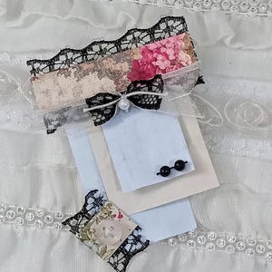 May include: A mini 3 pg. paper notepad featuring layered light blue and cream paper sheets. The top is decorated with black lace, a small black lace bow with a white pearl center, and pink floral accents. A small square with a clear button is attached.
