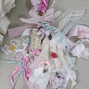 May include: A handmade decorative item with a variety of ribbons, lace, and fabric scraps in shades of pink, white, and blue. Embellishments include small roses, buttons, and charms. The word "Love" is visible.