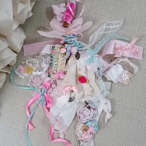 May include: A decorative fabric tassel ornament featuring layered ribbons, lace, and tulle in shades of pink, white, and blue. It includes small fabric flowers, buttons, and a rectangular tag with the word forever, topped with a teal love cutout.