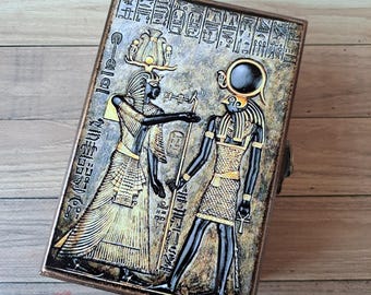 May include: A wooden box with a hinged lid featuring a detailed image of two Egyptian deities. The box is decorated with gold and black paint and features hieroglyphs. The deities are depicted in a standing position, with the female deity on the left and the male deity on the right. The female deity is wearing a crown and a long, flowing dress. The male deity is wearing a headdress and a short, pleated skirt. The box is a decorative item and would make a unique gift for anyone interested in Egyptian mythology.