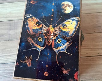 May include: A wooden box with a hinged lid featuring a colorful butterfly design against a dark blue background with a full moon and stars. The butterfly has yellow, orange, blue, and black wings.