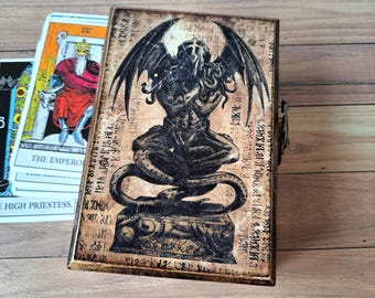 May include: A wooden tarot card box with a dark, antique finish. The box features a detailed black illustration of a winged, tentacled figure atop a pedestal, with ancient script-like text. Tarot cards are visible in the background.