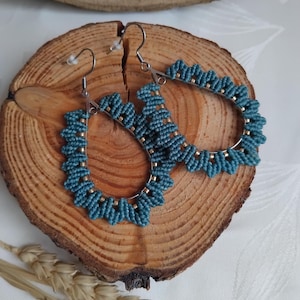 May include: A pair of teal blue beaded earrings with a teardrop shape and silver hooks. The earrings are displayed on a wooden surface.