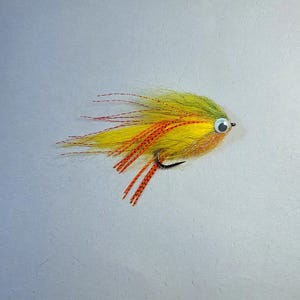 May include: A fishing fly lure featuring a bright yellow and olive green synthetic fiber body, accented with long orange and black speckled rubber legs, thin red tinsel strands, and a single prominent white and black googly eye on a metal hook.