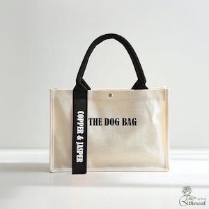 Op de afbeelding: Crèmekleurige draagtas met zwarte handvatten en een zwarte band. De tekst "THE DOG BAG" is op de voorkant gedrukt, met "COPPER & JASPER" verticaal op de band. De tas heeft een eenvoudig, strak ontwerp.