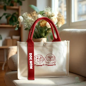 May include: Cream-colored tote bag with red handles and a red strap that says "DOG MOM." The bag features two dog illustrations and the text "COPPER ROCKET" in red. The bag is sitting on a beige couch.