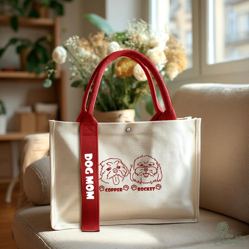 May include: Cream-colored tote bag with red handles and a red strap that says "DOG MOM." The bag features two dog illustrations and the text "COPPER ROCKET" in red. The bag is sitting on a beige couch.