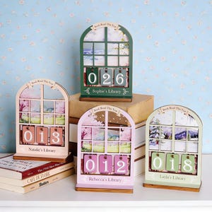 May include: Wooden book reading calendars in green, pink, and light green, each with a window design and a number display. The calendars have the text "Book Read This Year" and personalized names like "Sophie's Library". The numbers displayed are 026, 013, 012, and 018.