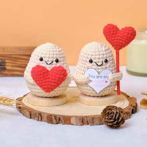 May include: Two crocheted potato-shaped figures with smiling faces. One holds a red heart, the other a sign that says "You are my sweet potato." Both are on wooden stands. A red heart on a stick is also present.