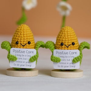 Könnte beinhalten: Zwei gehäkelte Maisfiguren mit lächelnden Gesichtern und grünen Armen, die Schilder mit der Aufschrift "Positive Corn" halten. Die Schilder enthalten den Text "I may be a tiny corn, but I believe in you. Go do your thing!" Sie stehen auf Holzsockeln.