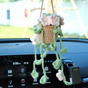 May include: A crocheted hanging plant with pink flowers and green leaves. The plant is in a brown pot and is hanging from a car's rearview mirror.