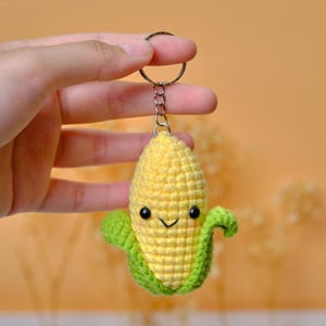 Könnte beinhalten: Ein gehäkelter Maiskolben-Schlüsselanhänger mit einem lächelnden Gesicht. Der Schlüsselanhänger hat einen gelben Maiskolben mit grünen Blättern und schwarzen Augen. Der Schlüsselanhänger wird von einem silbernen Metallring gehalten.