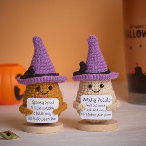 Puede incluir: Dos figuras de patata de ganchillo con sombreros de bruja morados. Una es de color canela y la otra es marrón claro. Cada una sostiene un letrero con texto temático de Halloween. Una calabaza y una taza con temática de Halloween están en el fondo.