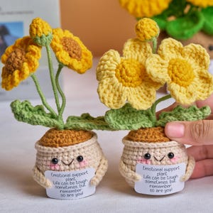 May include: Two handmade crochet sunflower plants in smiling pots. The yellow flowers have brown centers and green stems and leaves. Each pot has a smiling face and holds a sign that reads "Emotional support plant. Life can be tough sometimes, but remember so are you!"