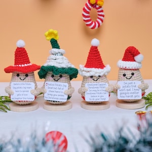 Op de afbeelding: Vier gehaakte aardappel figuren met feestelijke hoeden, elk met een bordje met een boodschap. De hoeden zijn rood, wit en groen, met een kleine kerstkrans op de achtergrond. De aardappelen staan op houten voetstukken.