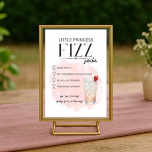 May include: A gold framed sign with the words "Little Princess Fizz Soda" and a drink recipe. The recipe includes Sprite, raspberry and peach syrup, creamer, and a raspberry garnish. The sign is on a pink tablecloth.