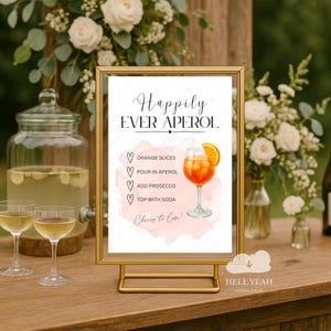 Può includere: Un cartello con cornice dorata con la scritta "Happily Ever Aperol" e una ricetta per cocktail. La ricetta include fette d'arancia, Aperol, Prosecco e soda. Il cartello è su un tavolo di legno con composizioni floreali.