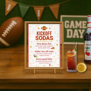 Könnte beinhalten: Ein gerahmtes "Kickoff Sodas"-Menü mit einem Football-Motiv, das Getränke-Rezepte auflistet. Ein Cocktail mit einer Zitronenscheibe und eine Flasche Kirschsirup stehen auf einem Holztisch. Ein Football und ein "Game Day"-Schild sind im Hintergrund.