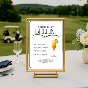 May include: A gold-coloured framed sign displaying a Bellini cocktail recipe. The sign includes instructions for the drink, with a golf course visible in the background. The sign is on a white table, with a wine glass and flowers nearby.