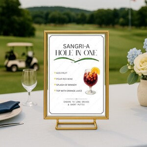 Può includere: Un cartello con cornice dorata con la scritta "SANGRI-A HOLE IN ONE" e una ricetta per la sangria. La ricetta prevede l'aggiunta di frutta, versare vino rosso, un goccio di brandy e guarnire con succo d'arancia. Il cartello è su un tavolo con un campo da golf sullo sfondo.