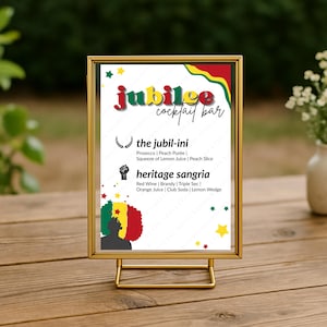 May include: A gold-framed sign for a "Jubilee Cocktail Bar" with a colorful design. The sign lists two cocktail recipes: "the jubil-ini" and "heritage sangria." The sign is displayed on a wooden table with a blurred green background.