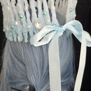 May include: A crown featuring tall, textured white icicle-like spikes adorned with small light blue pearls and clear faceted crystals. The base is secured with a light blue satin ribbon tied into a bow at the back, resting atop light blue hair.