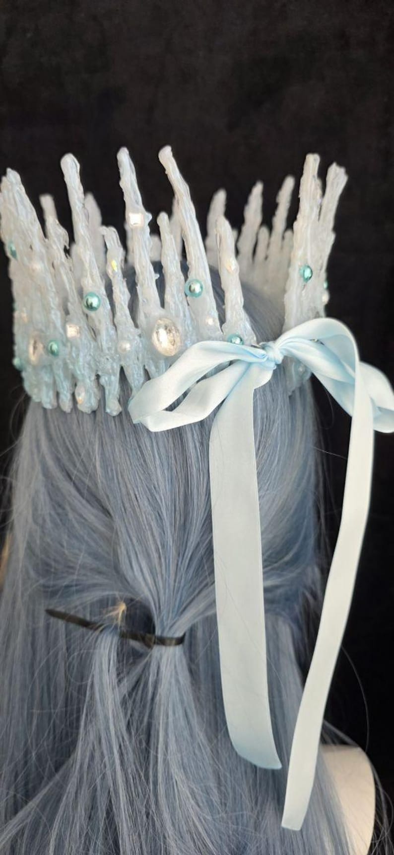 May include: A crown featuring tall, textured white icicle-like spikes adorned with small light blue pearls and clear faceted crystals. The base is secured with a light blue satin ribbon tied into a bow at the back, resting atop light blue hair.