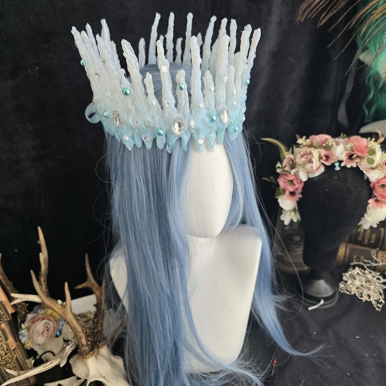 May include: A crown featuring tall, jagged, white and light blue icicle-like spikes. The base is adorned with small, round, light blue pearls and clear, oval-shaped faceted crystals that catch the light, creating a textured, crystalline appearance.