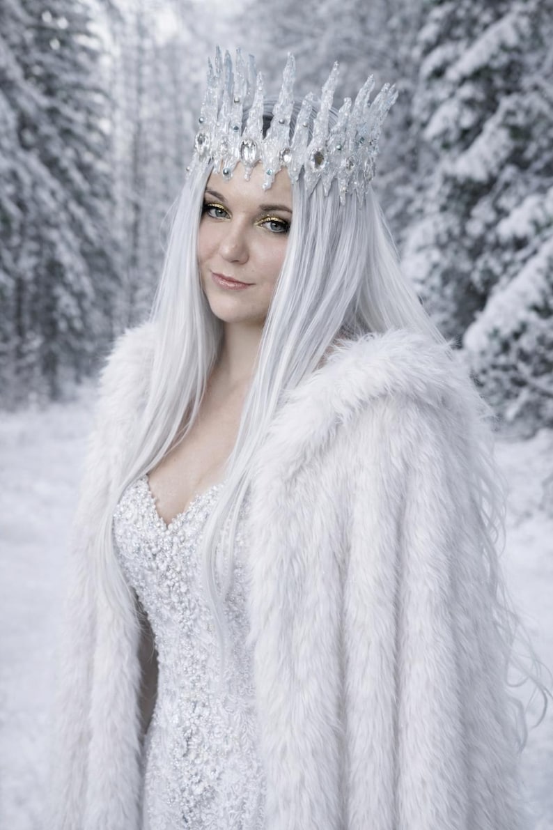 May include: A white faux fur cape worn over a heavily beaded, textured white gown. The ensemble includes a tall, crystalline crown featuring clear, pointed spikes and embedded oval-shaped gems, paired with long, straight white hair.