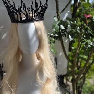 May include: A black crown featuring a jagged, branch-like structure with multiple pointed spikes extending upward. The crown is positioned on a white mannequin head that has long, wavy, light blonde hair flowing down past the shoulders.