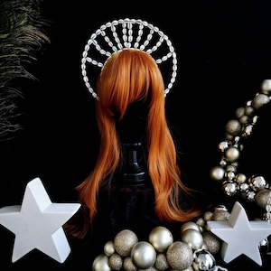 Può includere: Una corona di conchiglie bianche con un design circolare si trova su una parrucca rossa. La corona è fatta di piccole conchiglie bianche disposte in un motivo circolare. La parrucca è su una testa di manichino. Lo sfondo è nero con ornamenti argentati e dorati.