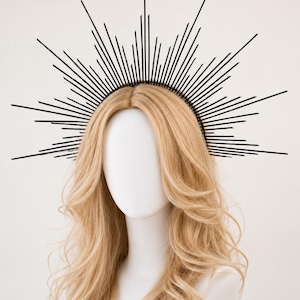 May include: A black sunburst crown headpiece featuring a series of thin, radiating spikes of varying lengths attached to a circular headband base. The spikes are arranged in a fan-like pattern, creating a sharp, geometric silhouette around the head.
