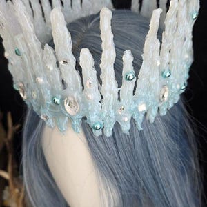 May include: A crown featuring tall, jagged, translucent white icicle-like spikes. The base is accented with light blue, iridescent, and clear circular beads and faceted crystals, creating a frozen, crystalline appearance on a mannequin head.