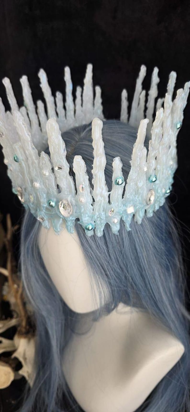 May include: A crown featuring tall, jagged, translucent white icicle-like spikes. The base is accented with light blue, iridescent, and clear circular beads and faceted crystals, creating a frozen, crystalline appearance on a mannequin head.