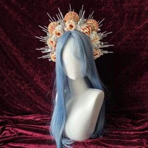 Può includere: Un copricapo decorativo con conchiglie, fiori finti e dettagli argentati. Il copricapo è esposto su una testa di manichino con lunghi capelli blu chiaro. Il design è intricato e ornato.