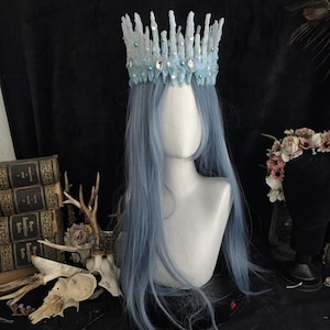 May include: A light blue crown with a jagged, ice-like texture, adorned with clear crystals and small teal beads. The crown is displayed on a mannequin head featuring long, straight, light blue hair, creating a striking and detailed headpiece.